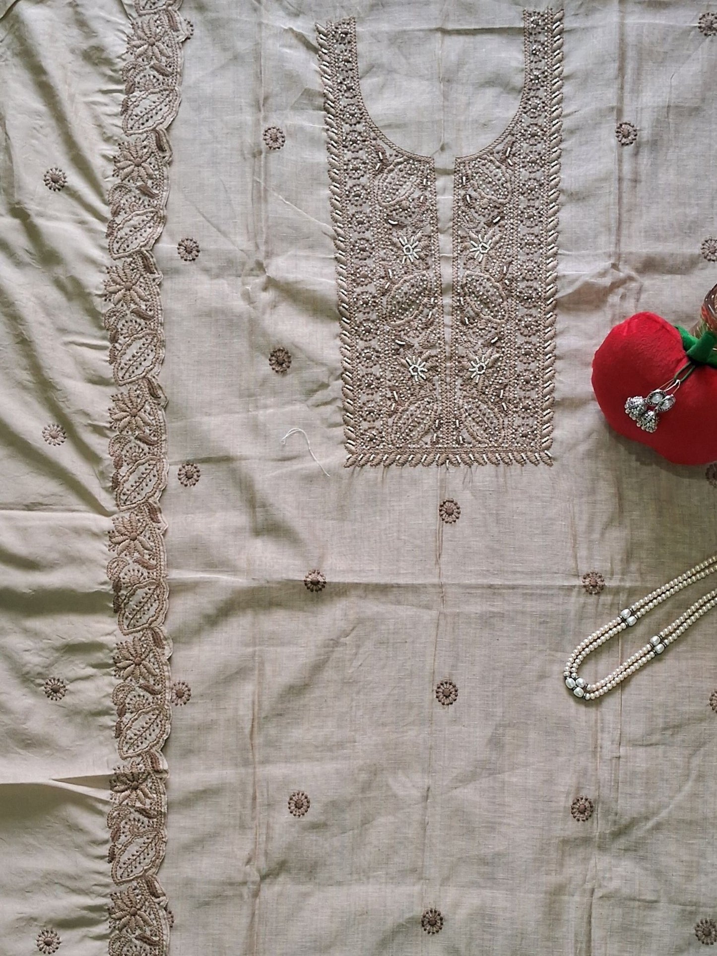 An ivor beige khadi chanderi embroidered unstitched salwar suit set laid flat, featuring a heavily embroidered neckline with floral motifs, a matching vertical border on a dupatta, and small embroidered dots across the fabric. A red decorative apple and a pearl necklace are styled as props on the right side.