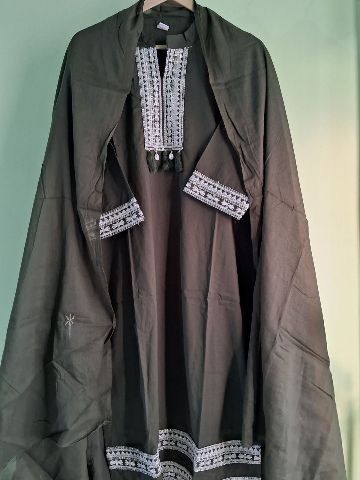 A mehendi green mul cotton ready to wear suit set with open-front tunic with white geometric embroidery on the neckline, sleeve cuffs, and hem, hanging against a pale green background.