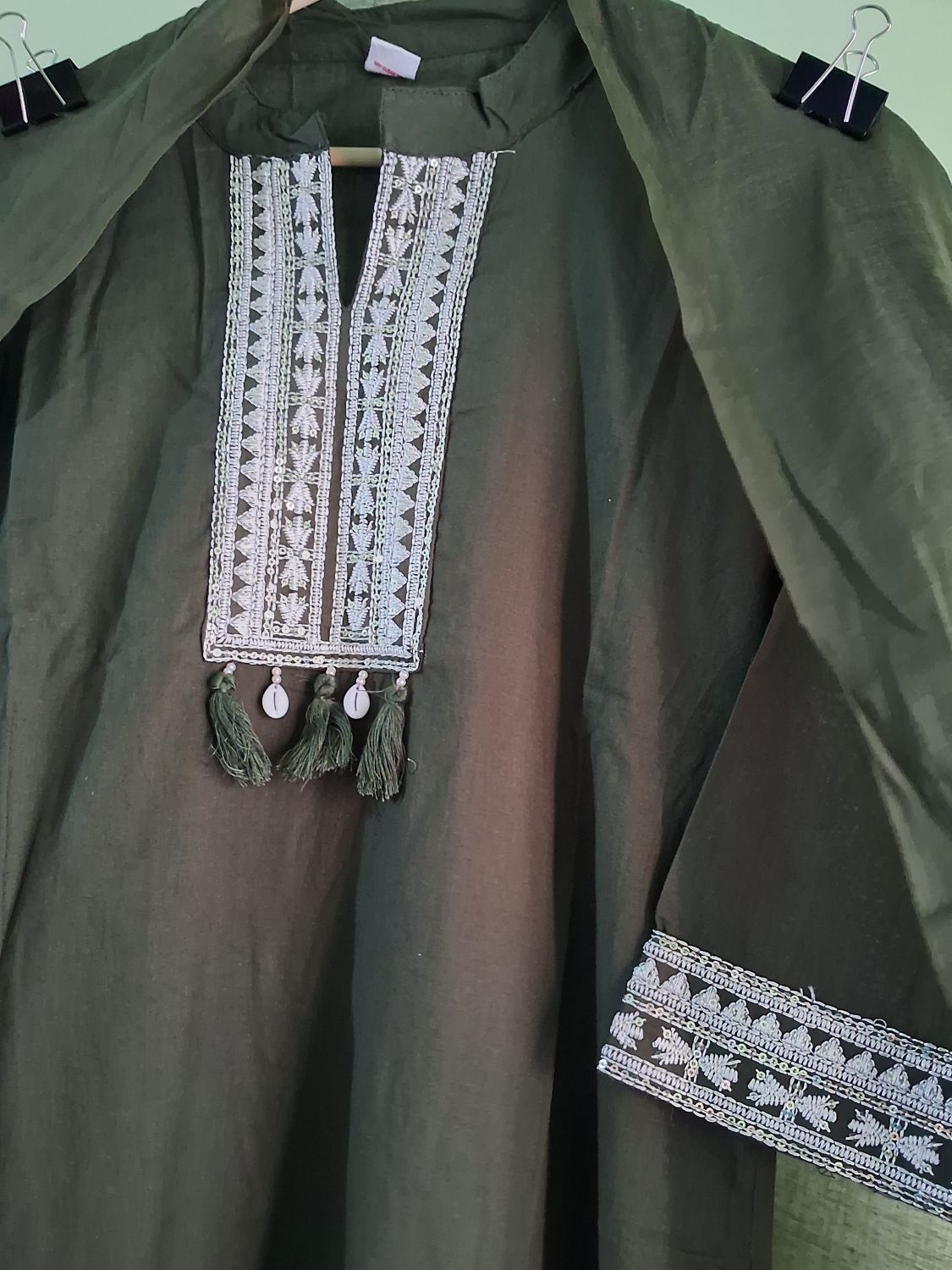 An olive green mul cotton ready to wear suit set with intricate white and silver embroidery on the chest panel and sleeve cuff, featuring hanging tassels and small white beads.