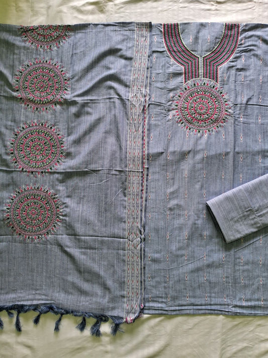 A flat-lay view of a light blue meerut cotton unstitched suit set with a matching dupatta, featuring pink and red embroidered circular mandalas on the dupatta and neckline, and a matching rolled-up sleeve.