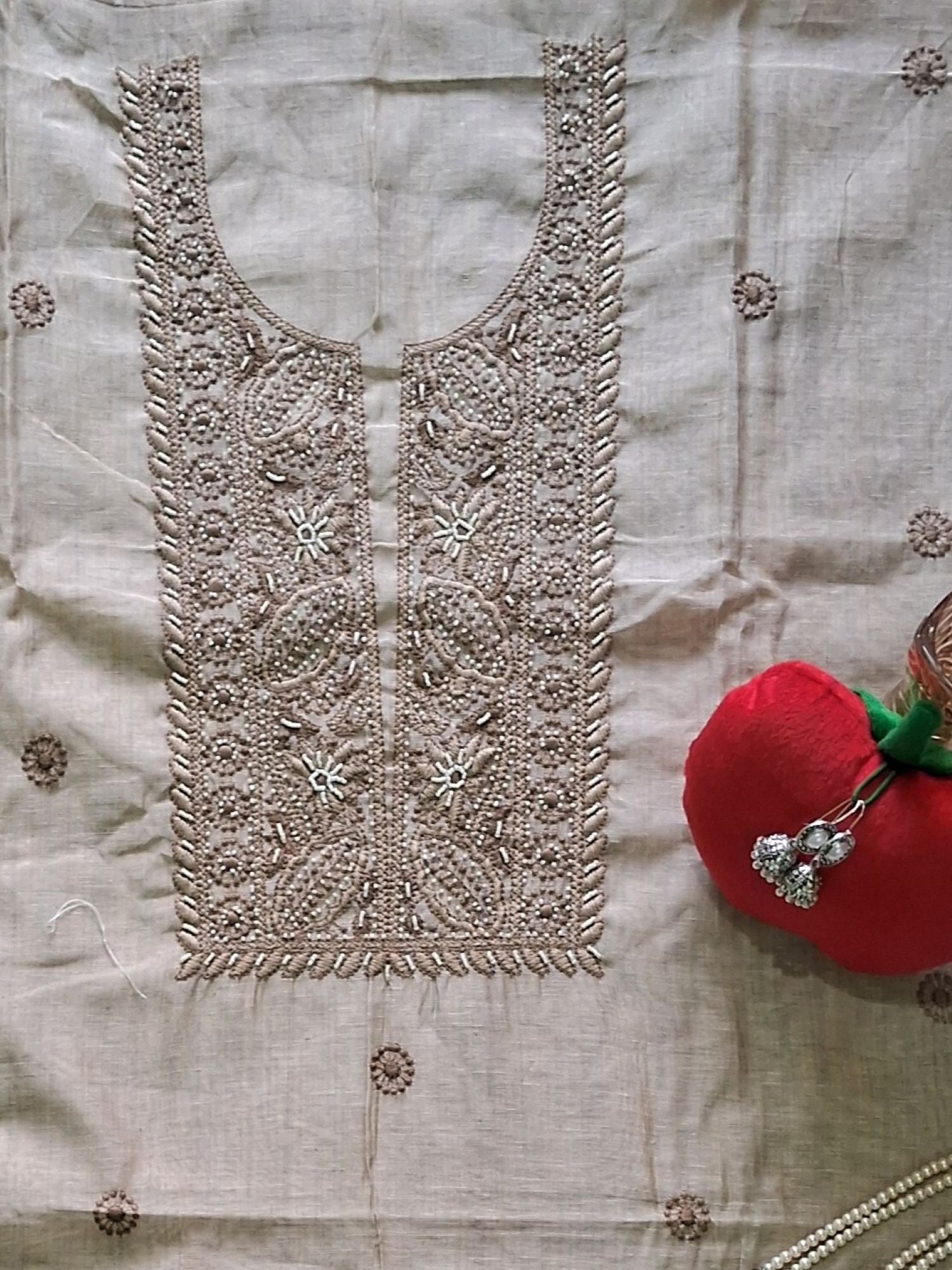An ivory beige khadi chanderi embroidered fabric panel with intricate floral and geometric stitching, laid flat on a matching beige textile background. A red pincushion with silver rings is placed to the right as a styling prop.