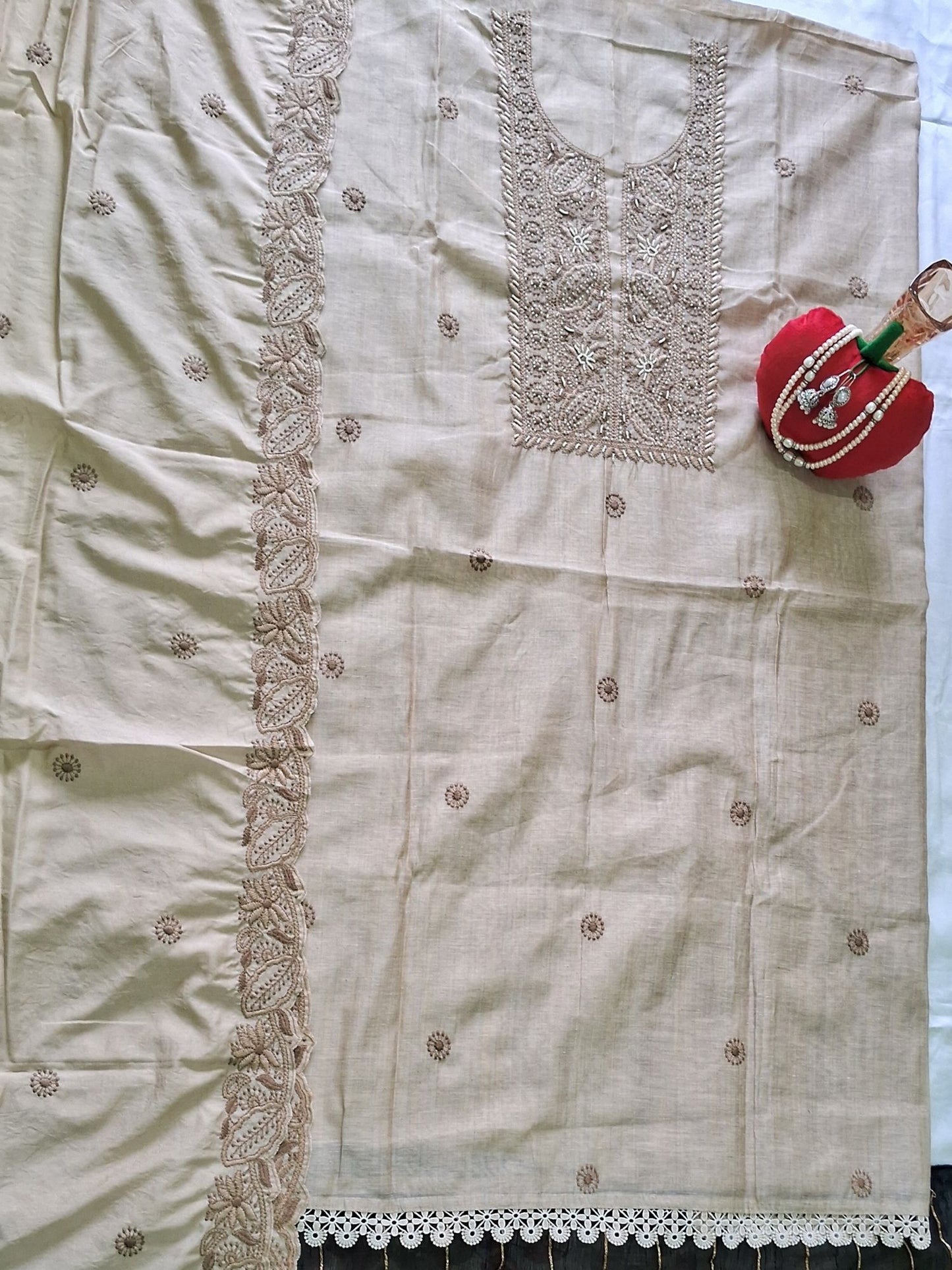 A light ivory beige khadi chanderi fabric set, likely a salwar suit material, featuring a kurta with detailed embroidery on the chest, a matching embroidered border, and a decorative dupatta draped over the side. A small red and green decorative pouch is placed on the upper right.