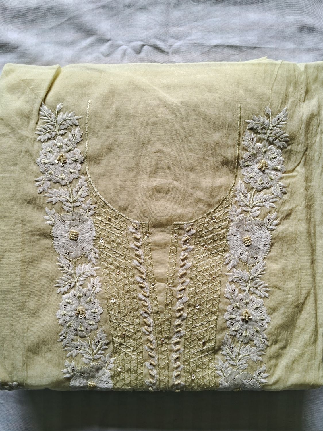 A folded light yellow unstitched kurta featuring intricate white floral embroidery and metallic gold thread detailing around the neckline, laid flat on a gray fabric background.