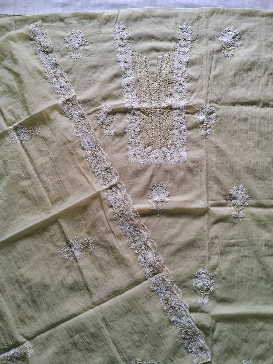 A light lemon yellow mul chanderi fabric, unstitched suit flat laid with top, bottom and dupatta featuring intricate white floral embroidery and lace borders.