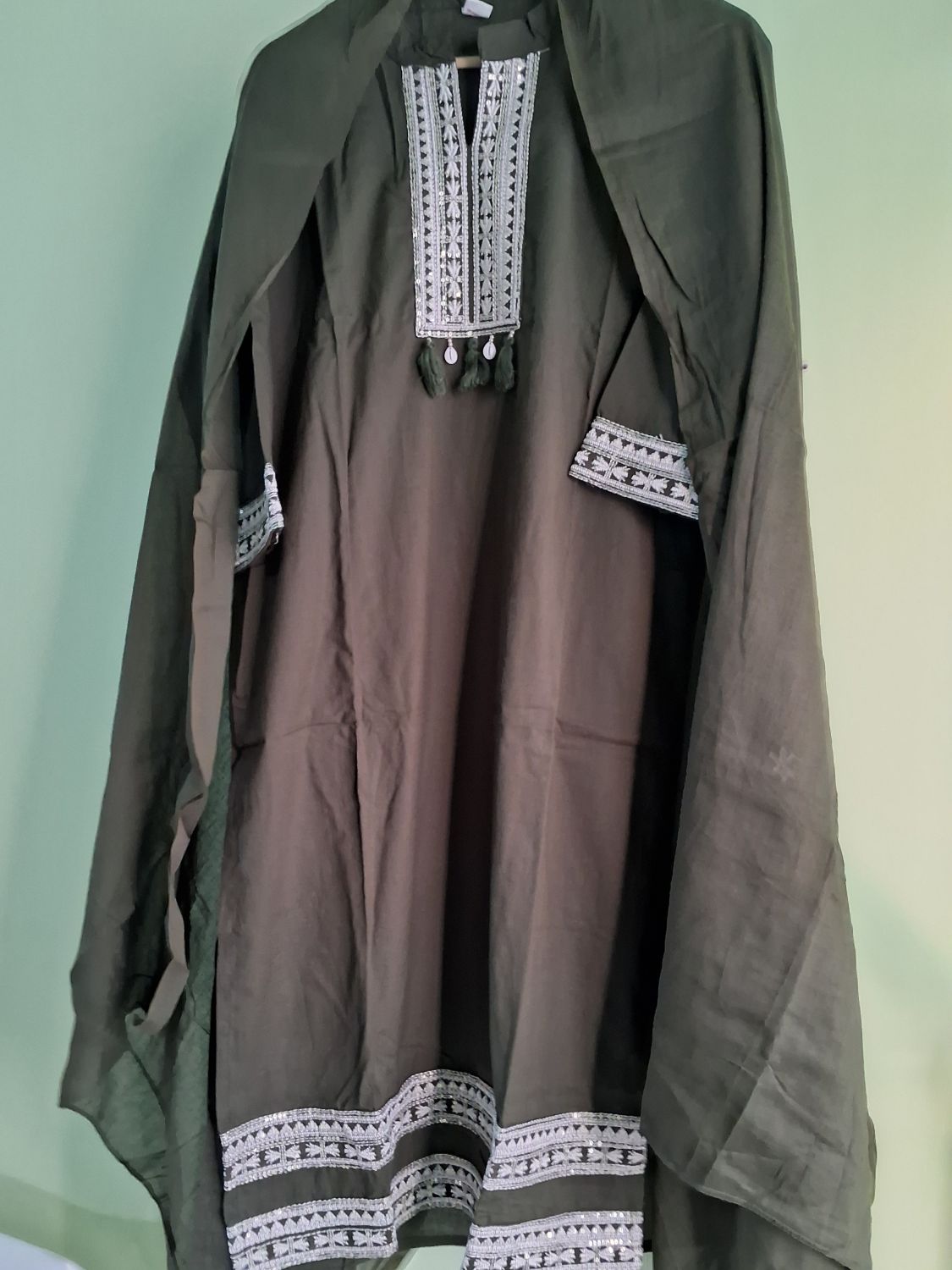 An olive green mul cotton ready to wear suit set  with white geometric embroidery on the neckline, sleeve cuffs, and hem, hanging against a light green wall.