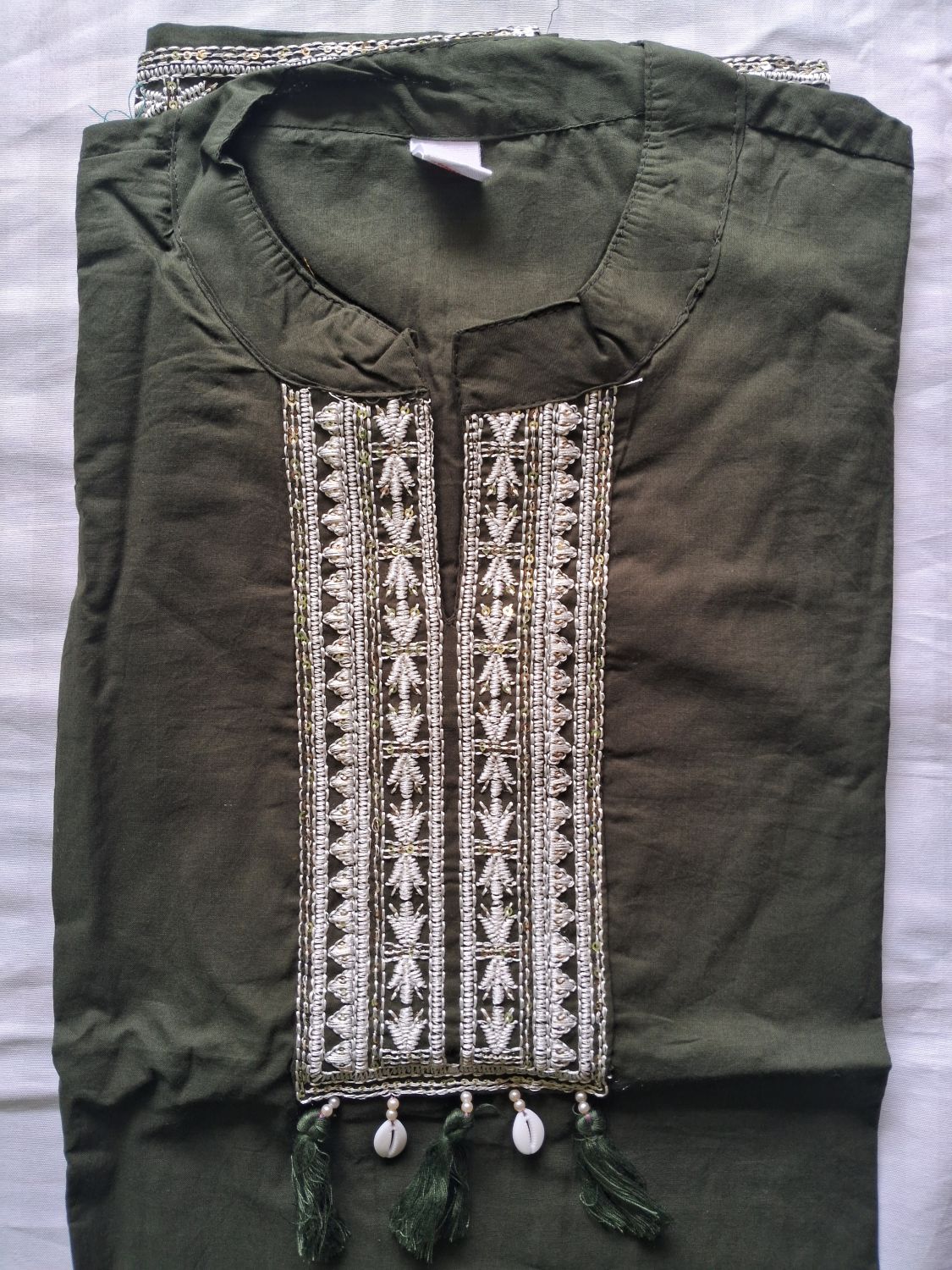 An olive green mul cotton ready to wear suit set folded cotton tunic top featuring a detailed vertical panel of white and silver geometric embroidery with tassel accents at the neckline, laid flat on a white fabric background.
