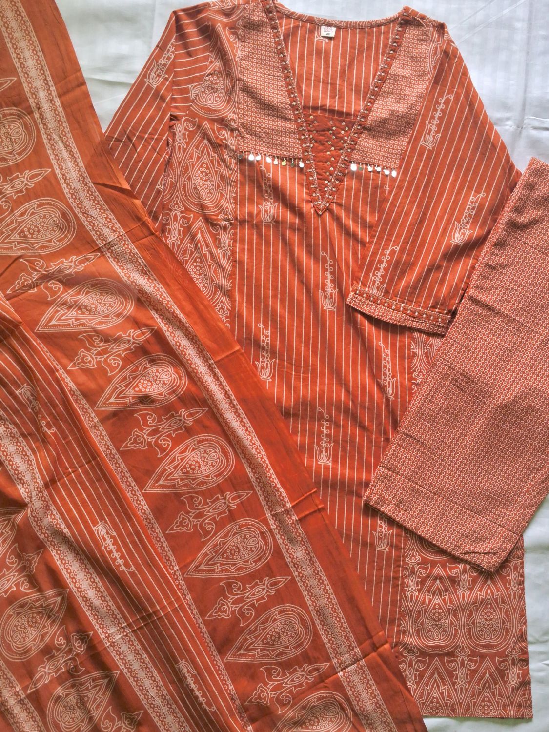 An orange and white printed three-piece salwar kameez suit set laid flat on a light gray fabric background. The set includes a long-sleeve tunic with vertical stripes and embroidery, matching trousers, and a dupatta with paisley and geometric patterns.