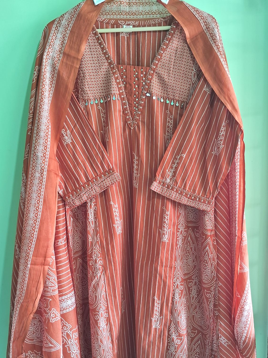 A rust colored printed cotton  traditional outfit consisting of a long-sleeved kurta with vertical stripes and white embroidery, paired with a matching sheer dupatta draped over the shoulders, hanging against a mint green background.
