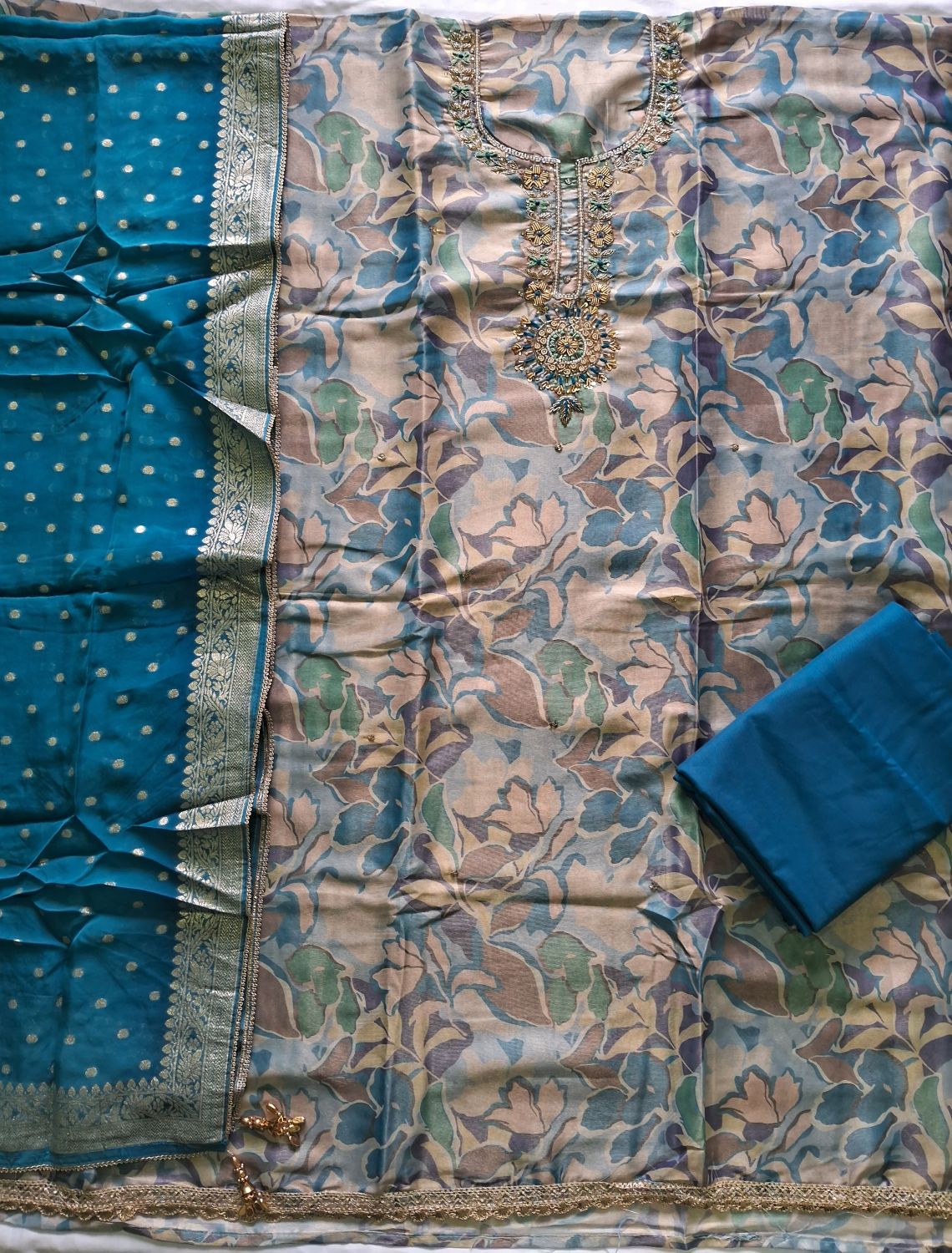 A traditional Indian salwar suit set fabric shimmer organza laid flat, featuring a floral print kurta in muted blue, beige, and green tones, a solid teal blue banarasi dupatta with silver zari borders, and a folded teal blue bottom piece.