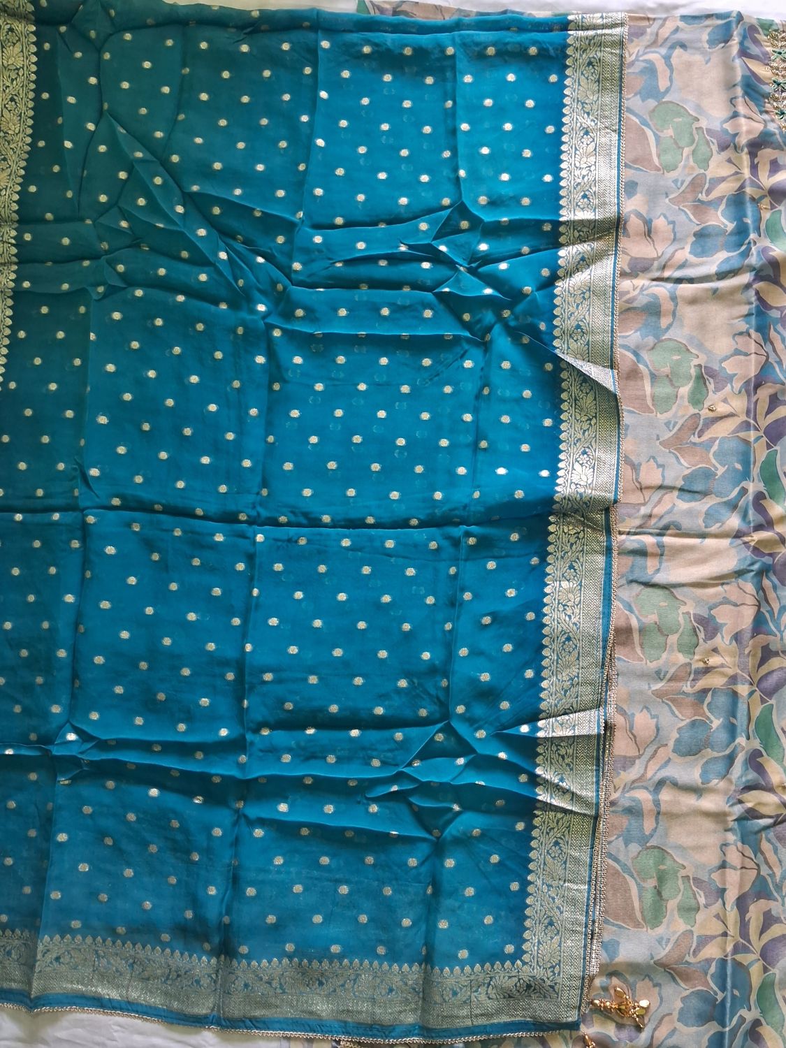 A teal blue shimmer organza unstitched suit with banarasi dupatta and small white polka dots and a wide beige border featuring a floral pattern in muted green, purple, and blue, with a metallic gold zari border on the top.