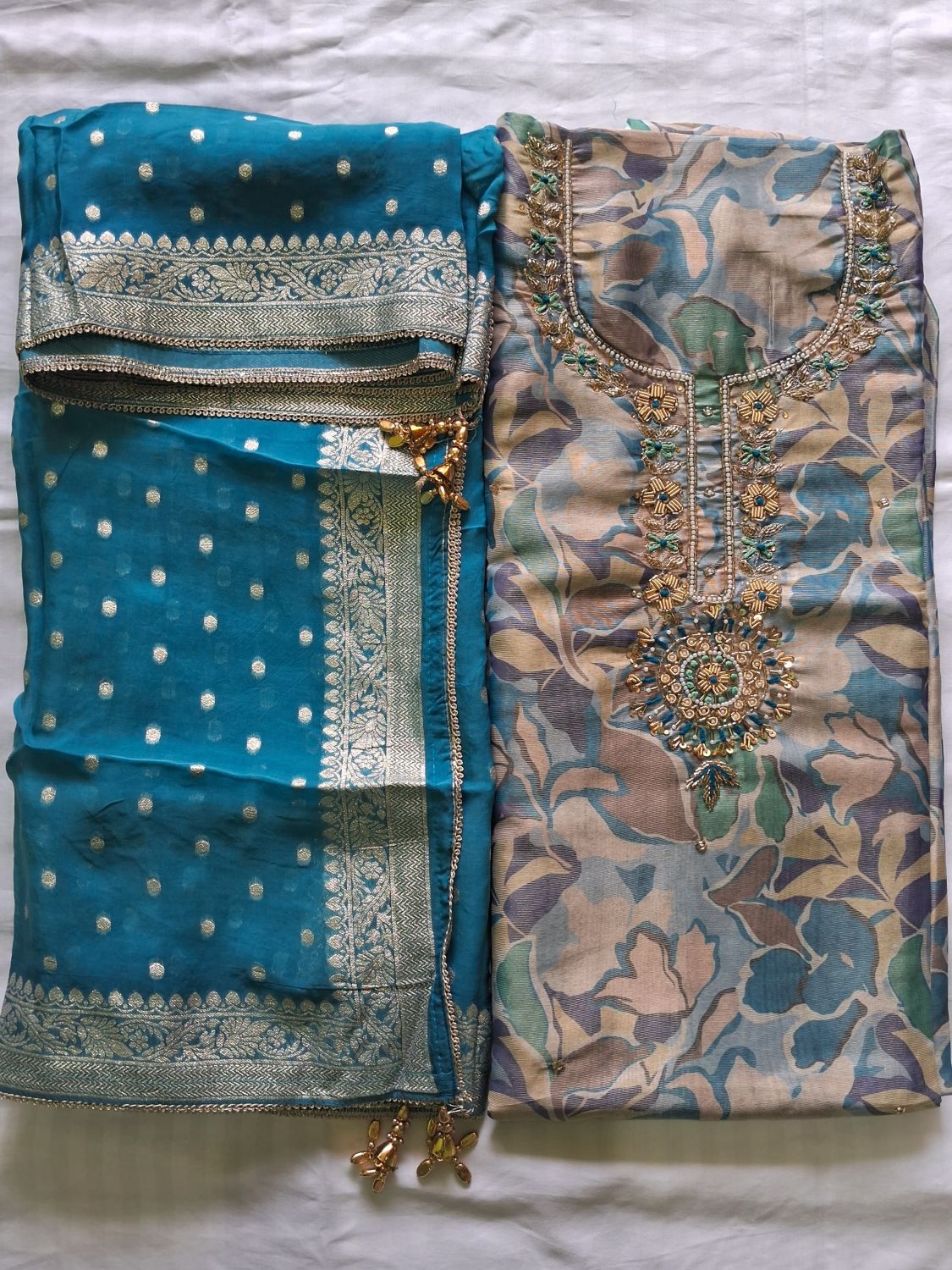 A folded traditional Indian salwar kameez suit set laid flat on a white fabric background. The set includes a teal blue printed kurta with gold and silver embroidery, a matching teal blue dupatta with white polka dots and a silver border, and a beige floral printed kameez shirt with a large gold and green embroidered neckline.