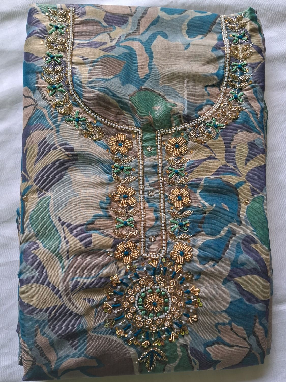 A teal blue folded women's kurta of shimmer organza fabric with an abstract floral print in muted blue, green, beige, and gray tones, featuring ornate gold and turquoise embroidery around the neckline and a large matching embroidered motif at the center front.