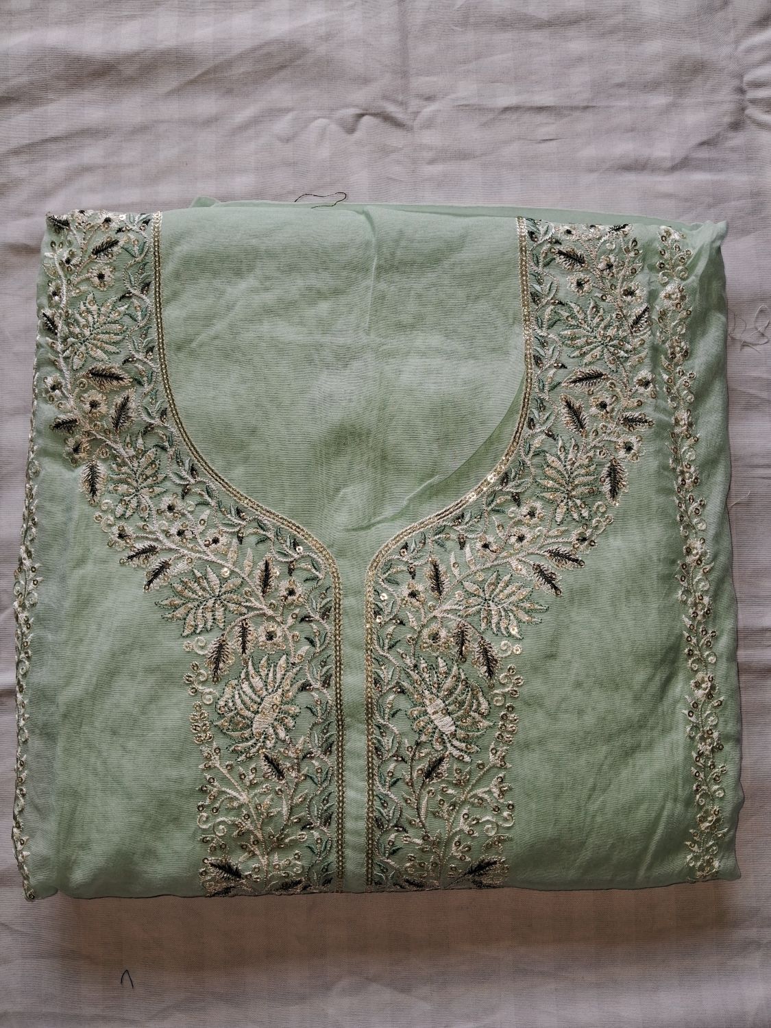A folded powder blue mul chanderi fabric unstitched suit, likely a traditional kurta or dress, featuring intricate floral embroidery and beadwork around the neckline and front border, laid flat on a neutral fabric background.