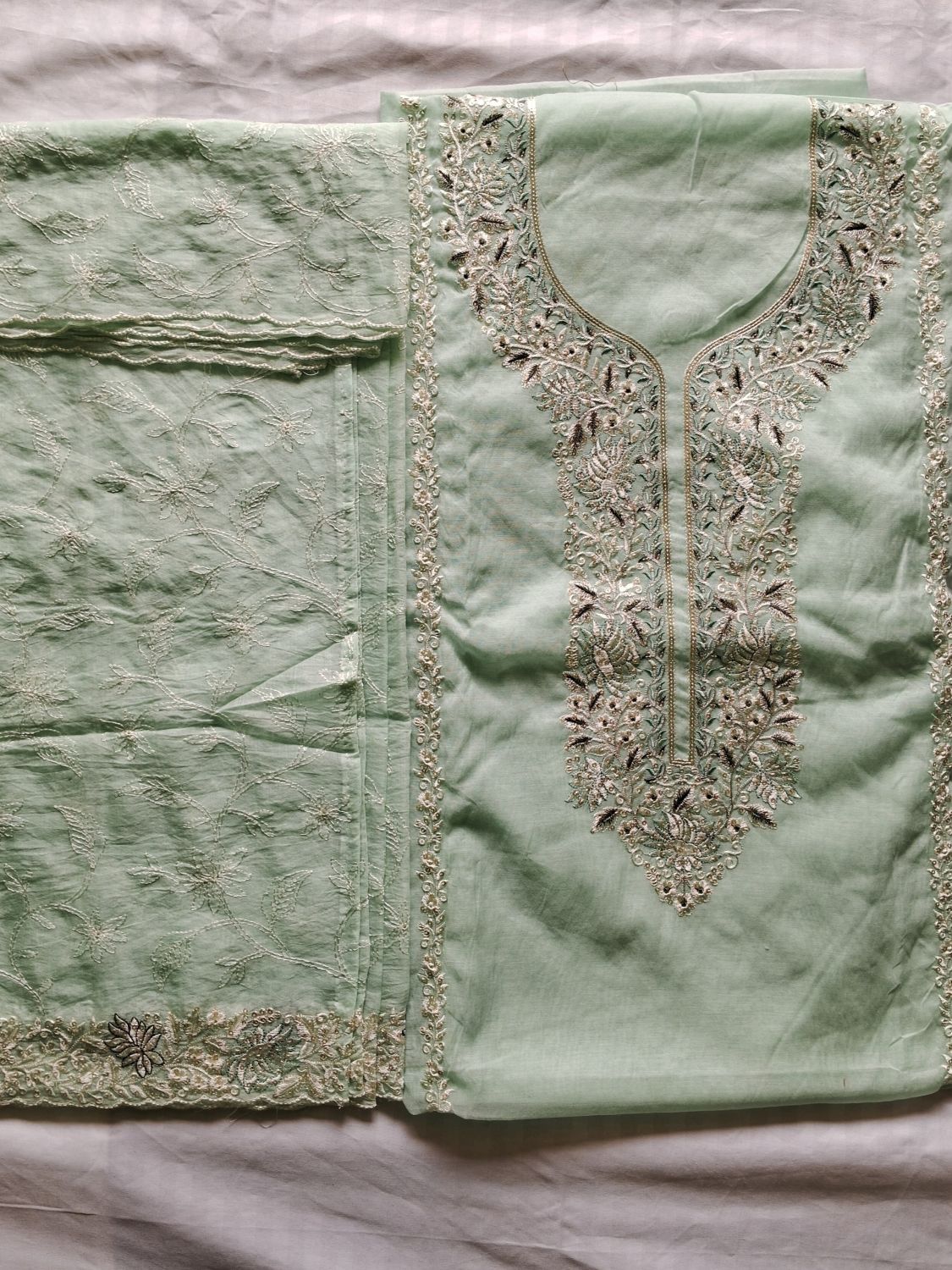 A light powder blue embroidered mul chanderi fabric unstitched suit set laid flat on a white fabric background, featuring a kurta-style top with ornate metallic embroidery around the neckline and a matching embroidered lucknowi chikankari dupatta.