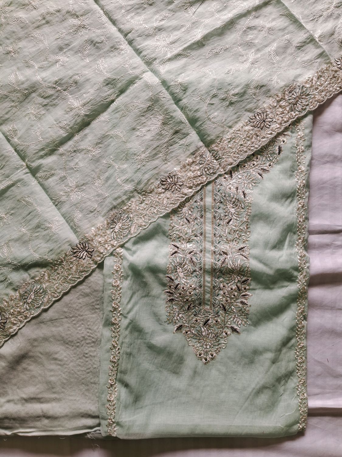 A close-up of a light powder blue unstitched dress material of mul chanderi fabric laid flat, featuring a matching embroidered lucknowi chikankari dupatta and border with metallic beige detailing.
