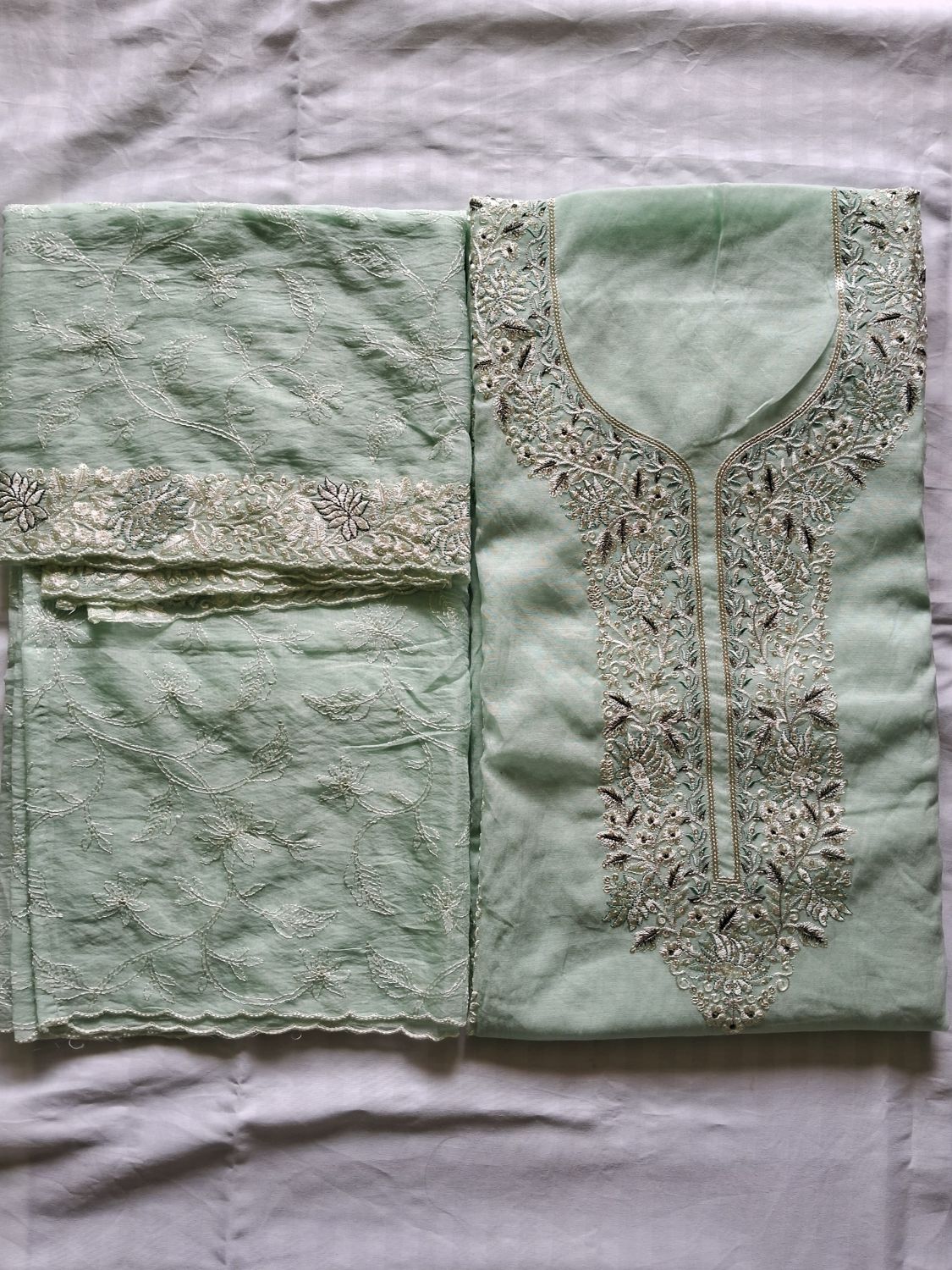 A folded mint green salwar suit material set with intricate silver and white embroidery, laid flat on a white fabric background.