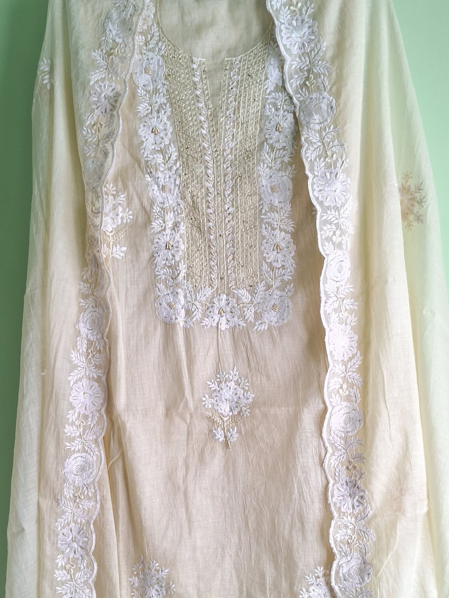 A cream-colored traditional embroidered unstitched dress of mul chanderi fabric with intricate white floral lace embroidery along the neckline, sleeves, and border, set against a light green background.