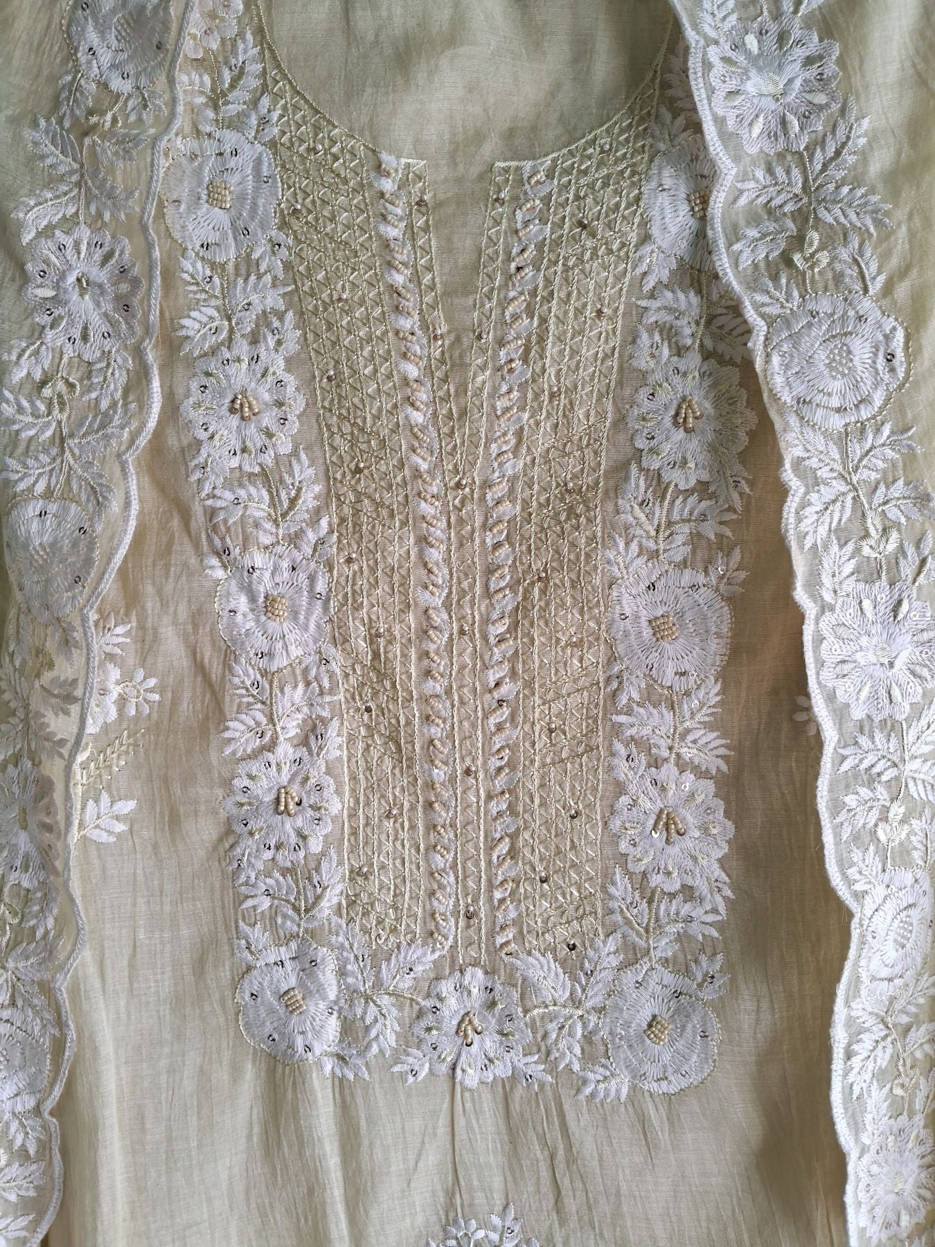 Close-up of a lemon yellow or beige -colored embroidered mul chanderi  fabric, showing detailed white floral lace embroidery and stitched trim around the neckline and front panel.